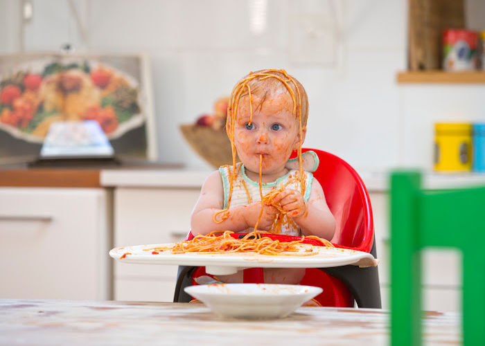 little baby in highchair eating pasta and covered in pasta