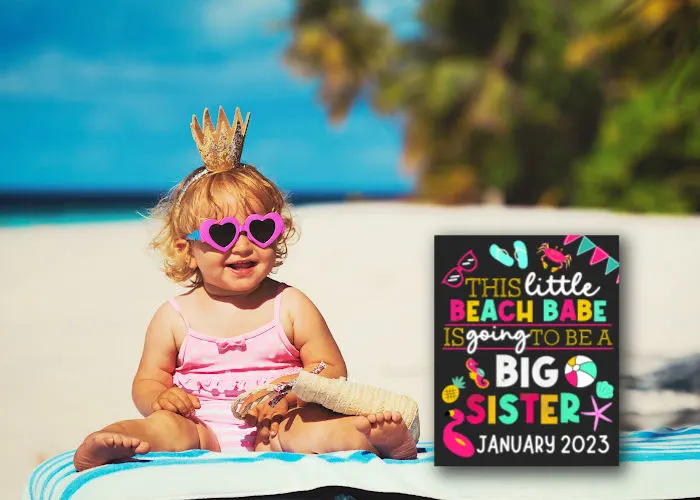 cute toddler girl in sun glasses at the beach sitting next to beach big sister announcement board