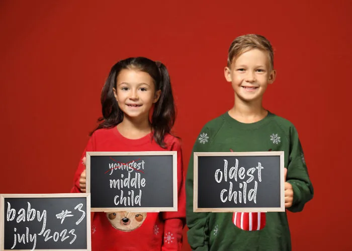 brother and sister dressed in Christmas sweater holding signs reading oldest child, middle child (with youngest crossed out) and baby #3 due July 2023