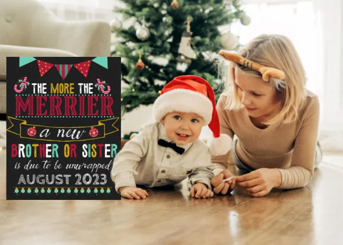 toddler boy in Santa hat with sister beside him as they lay down in front of Christmas tree next to the more the merrier Christmas pregnancy announcement sign