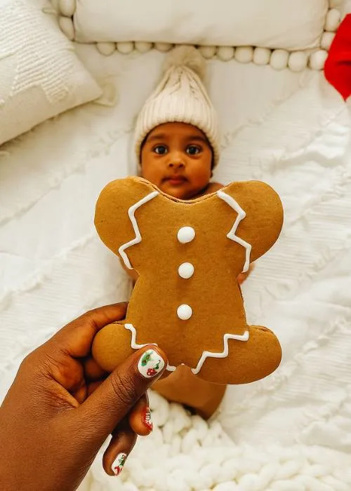 a mom's hand is holding a gingerbread cookie with the head bitten off in front of her baby. it creates the illusion of a baby gingerbread man