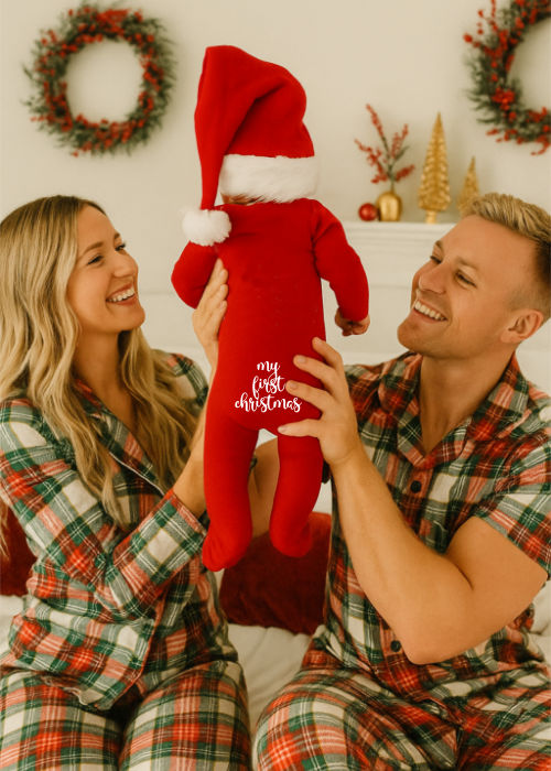 mom and dad are wearing their christmas pajama and holding up baby who has a onesie that says my first christmas