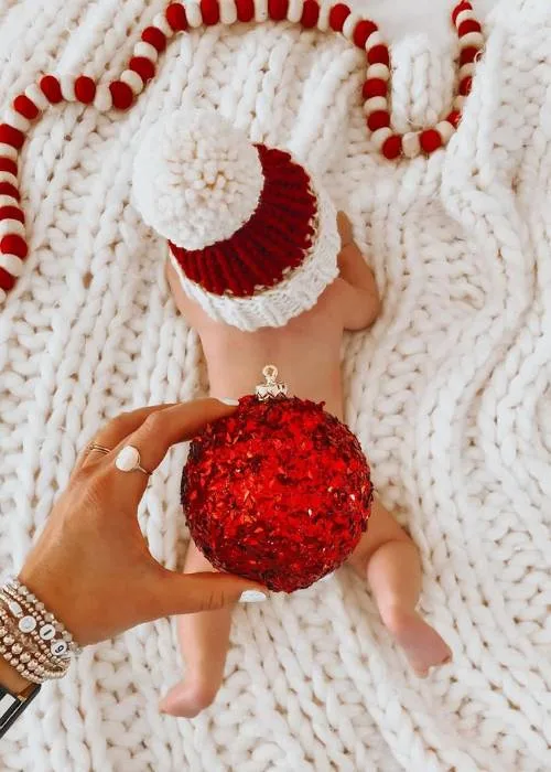 cute baby lying on knit blanket. a hand is holding a red ornament over baby's bum
