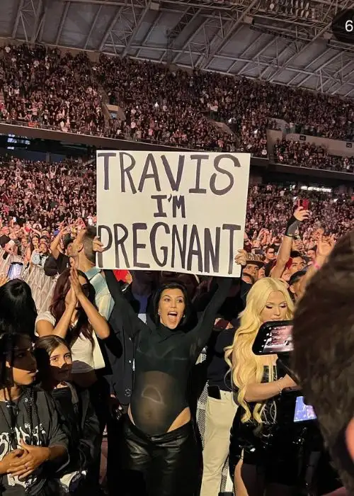 pregnant Kourtney Kardashian holding a sign that says "Travis I'm Pregnant'